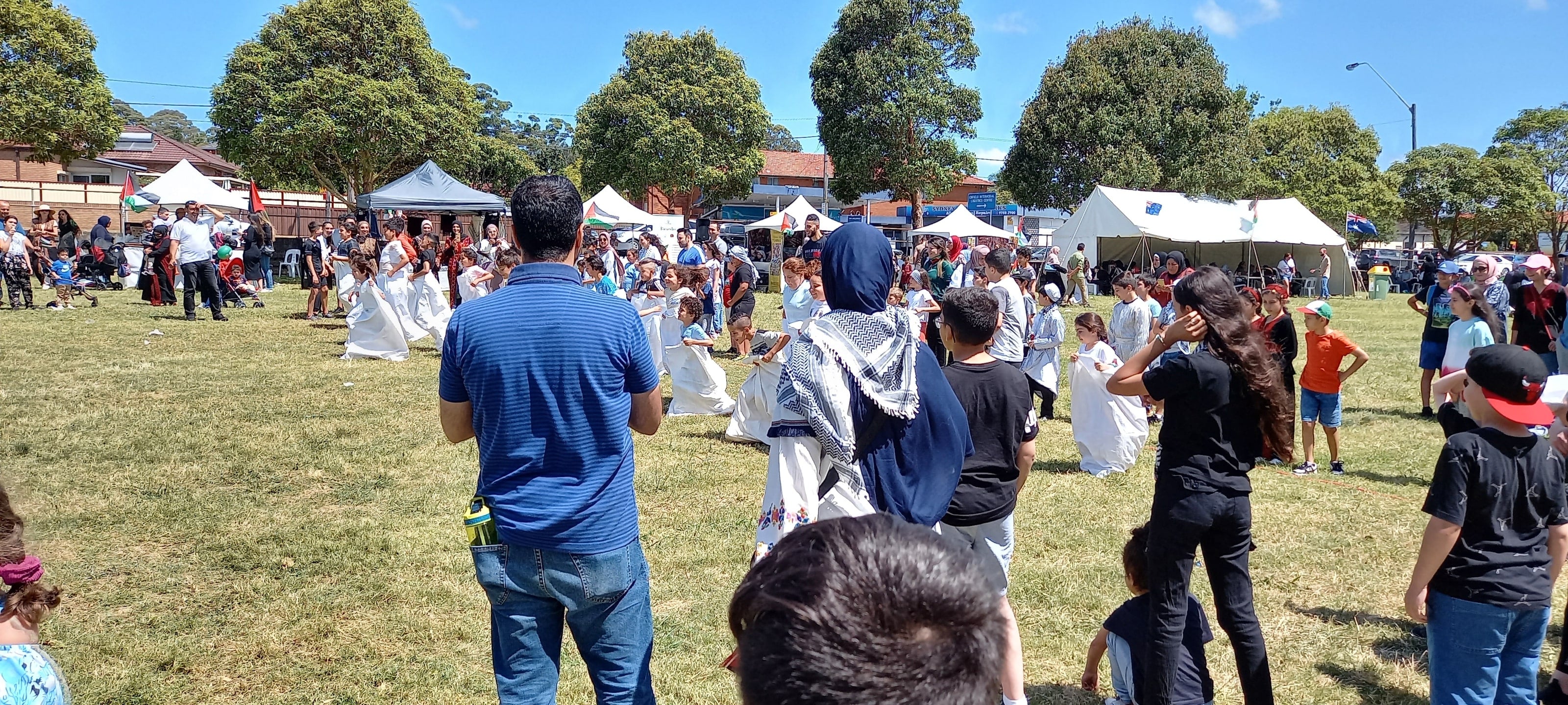 Families cheering on children in the sack race, a woman in a keffiyeh scarf in the middle